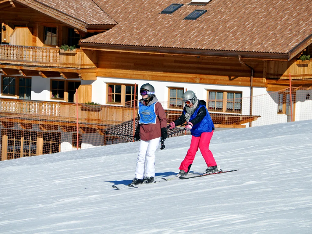 Zwei Skifahrende auf der Piste vor einem Holzhaus.